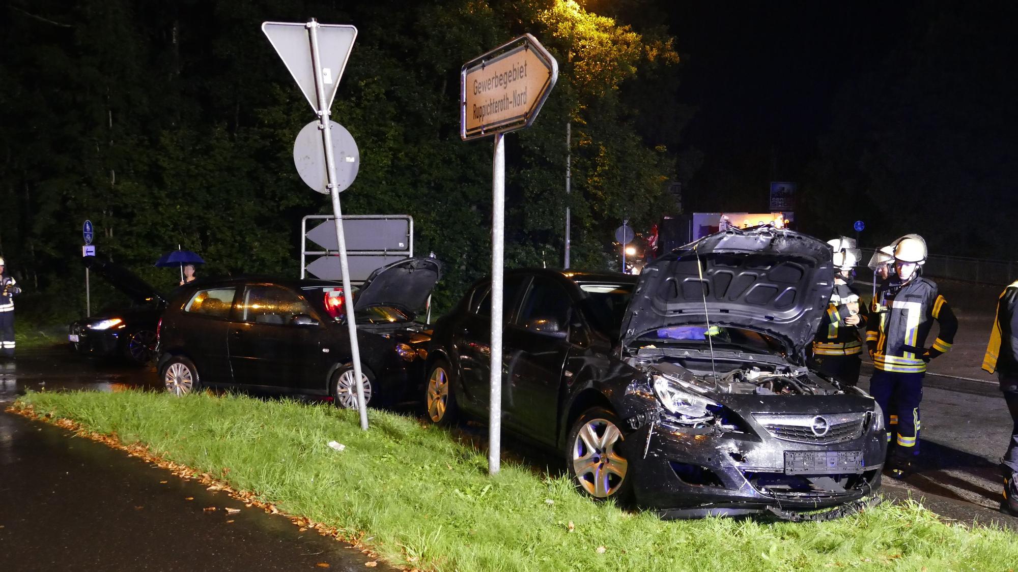 Ein beschädigtes Auto steht nach einem Unfall neben einem Verkehrsschild. Es ist Nacht und es regnet, Feuerwehrleute stehen auf der Straße.
