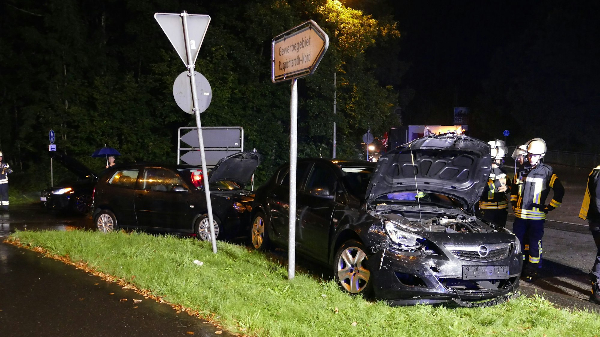 Ein beschädigtes Auto steht nach einem Unfall neben einem Verkehrsschild. Es ist Nacht und es regnet, Feuerwehrleute stehen auf der Straße.