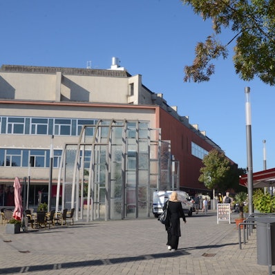 Ein großes Gebäude am Rand einer Fußgängerzone. Am Rand einer gepflasterten Fläche stehen Laternen und einige Bäume.