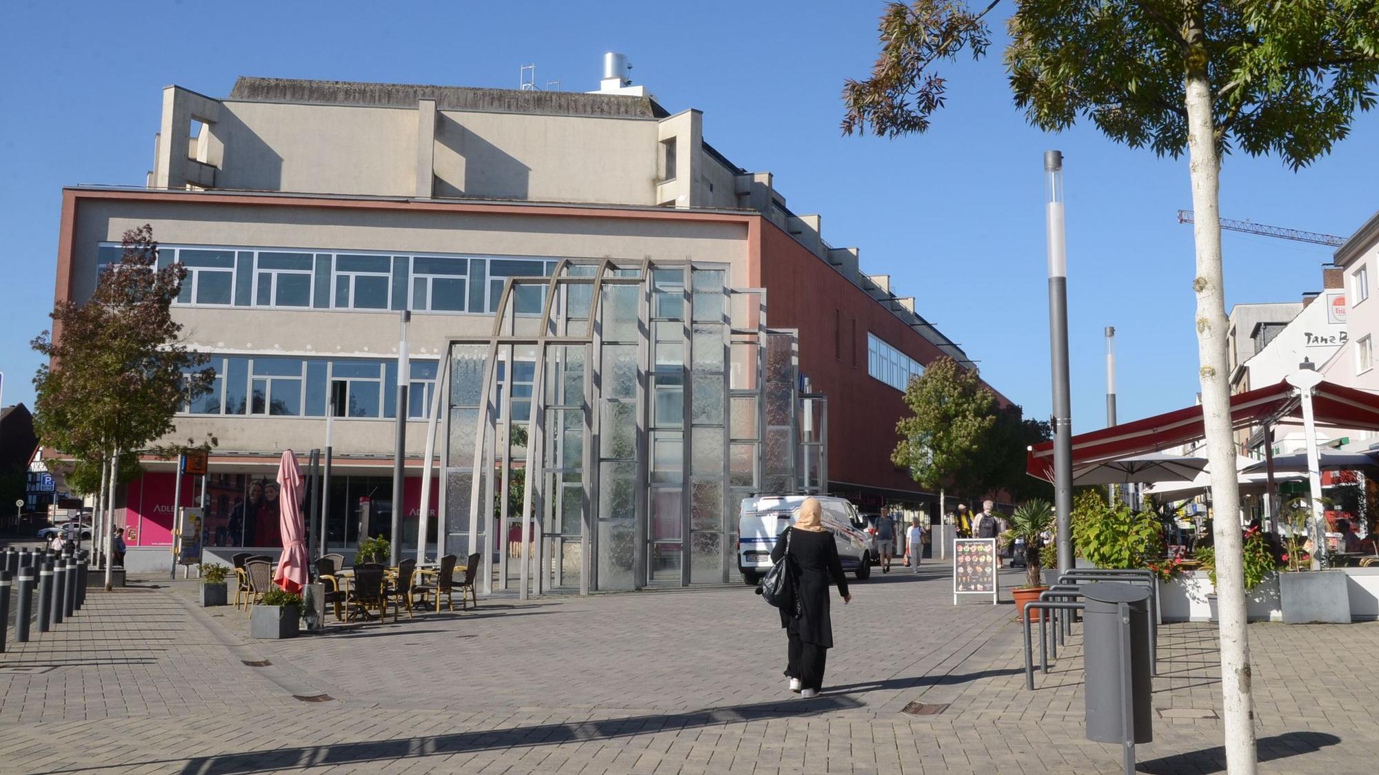 Ein großes Gebäude am Rand einer Fußgängerzone. Am Rand einer gepflasterten Fläche stehen Laternen und einige Bäume.