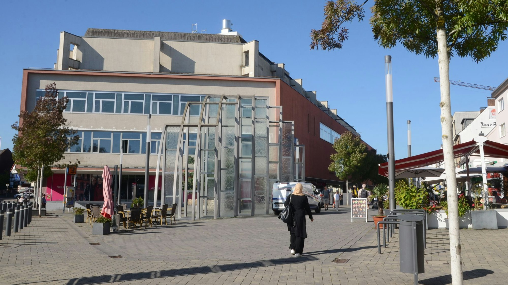 Ein großes Gebäude am Rand einer Fußgängerzone. Am Rand einer gepflasterten Fläche stehen Laternen und einige Bäume.