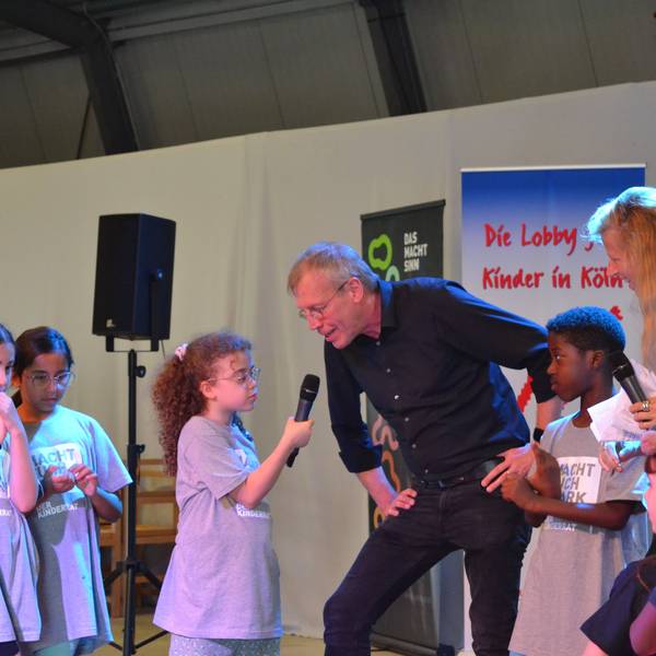 Kinder stehen zusammen und ein Kind hält einem Mann ein Mikrofon an den Mund.