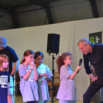 Kinder stehen zusammen und ein Kind hält einem Mann ein Mikrofon an den Mund.