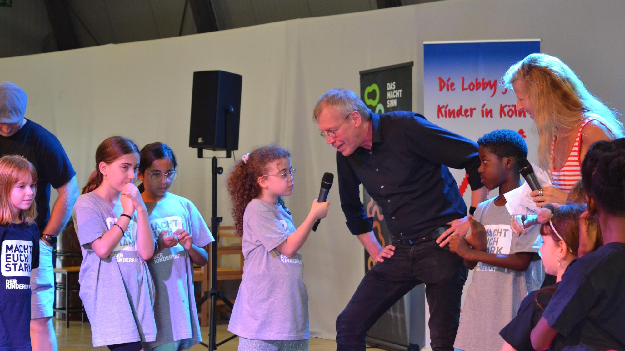 Kinder stehen zusammen und ein Kind hält einem Mann ein Mikrofon an den Mund.