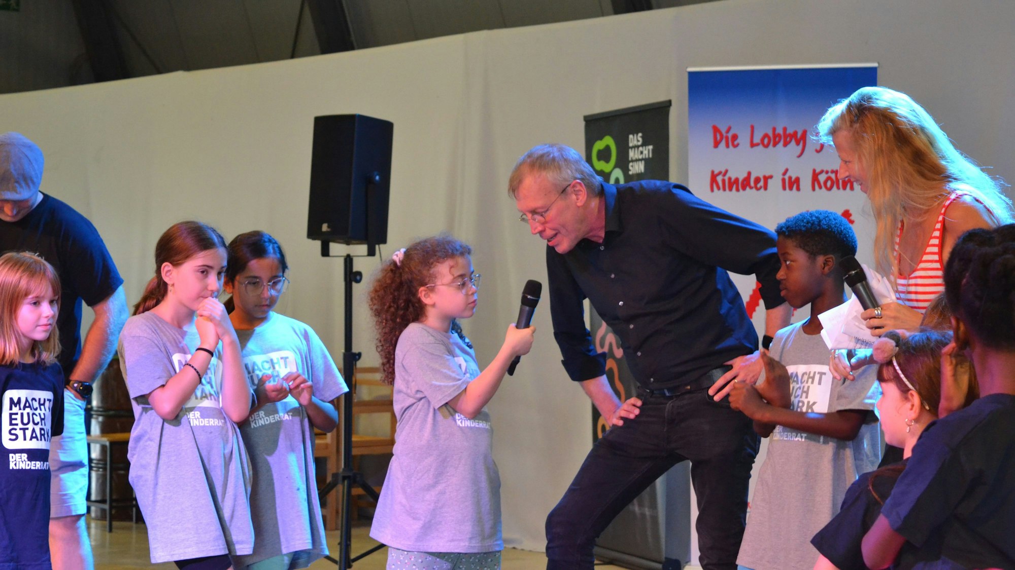 Kinder stehen zusammen und ein Kind hält einem Mann ein Mikrofon an den Mund.