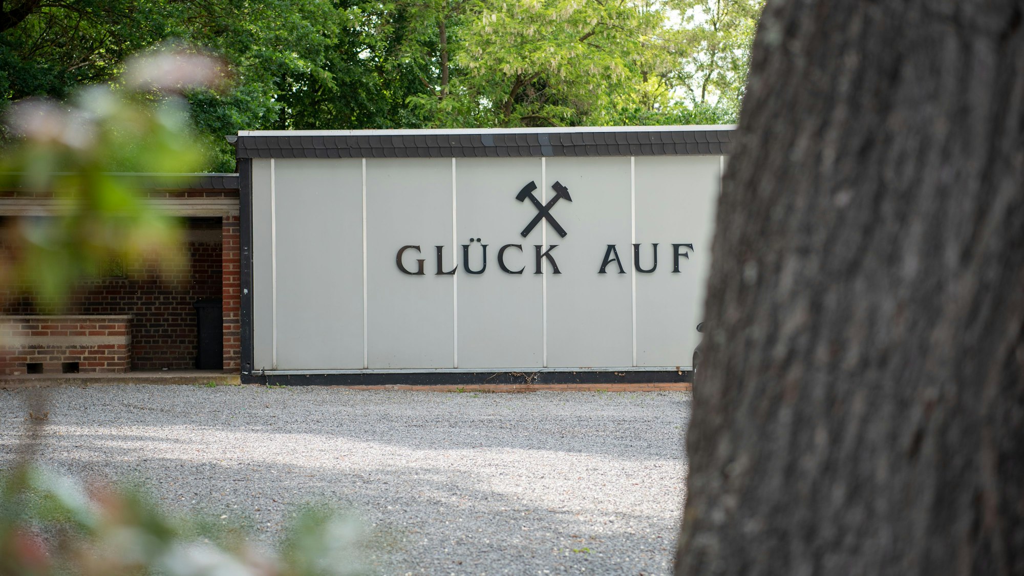 Auf einer Wand steht in großen Buchstaben „Glück Auf“. Davor ist ein Platz aus Kies, im Hintergrund sind Bäume zu erkennen.