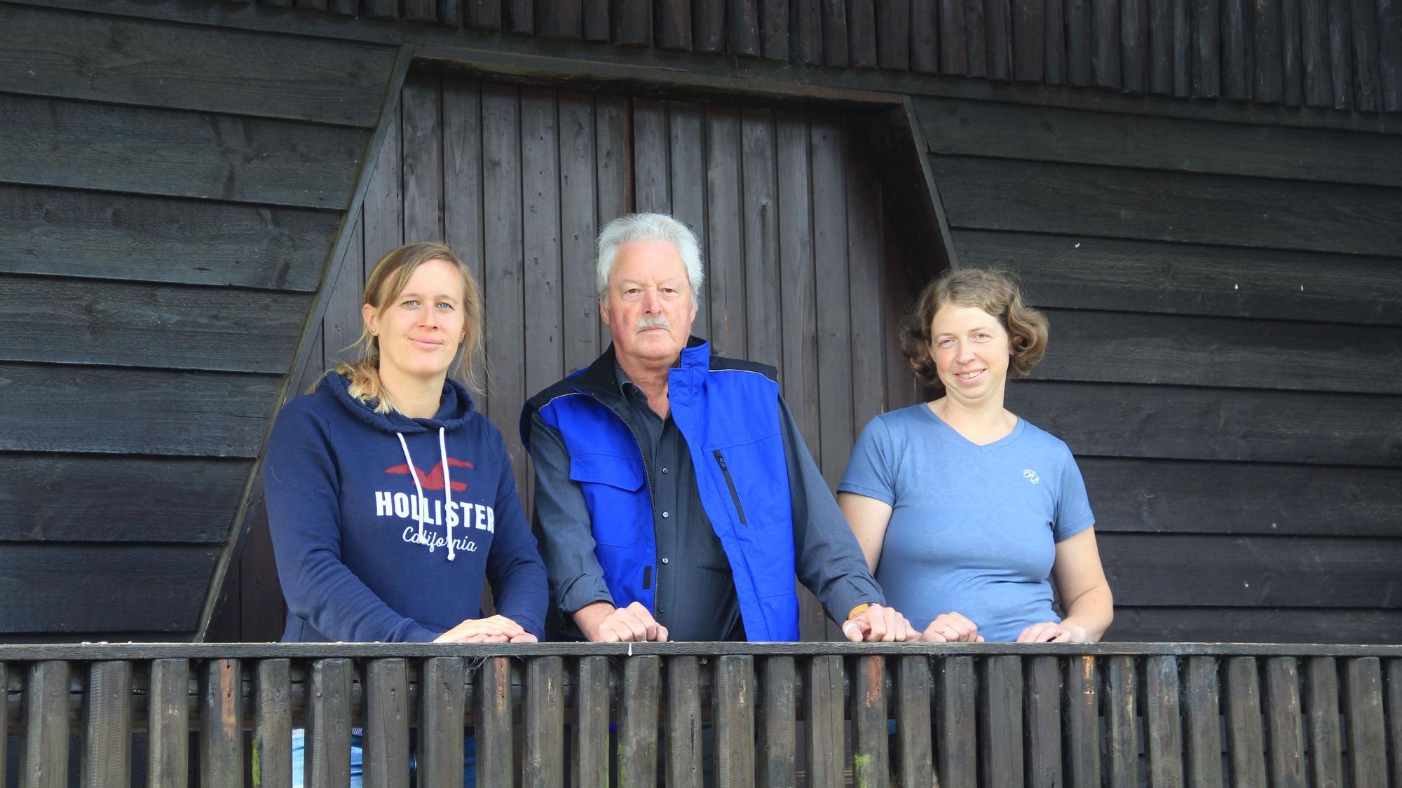 Ein Mann und zwei Frauen stehen auf einem Balkon eines Holzhauses.