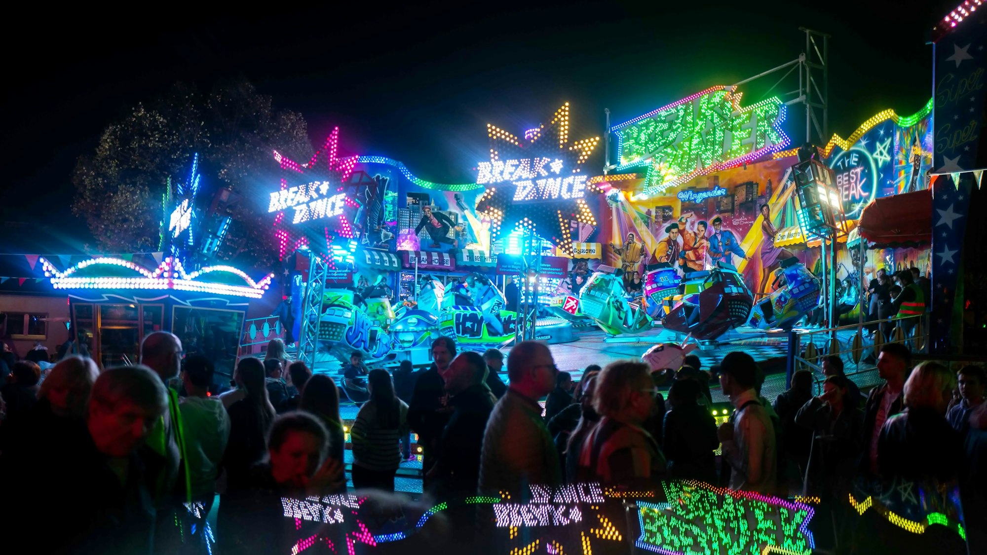Fahrgeschäfte auf der Kirmes erstrahlen in bunten Neon-Lichtern. Im Vordergrund schlendern Menschen über die Kirmes.