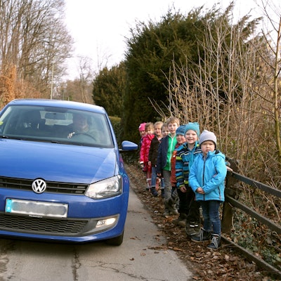 Ein blaues Auto fährt zwischen parkenden Pkw und einer Gruppe von kleinen Kindern hindurch.