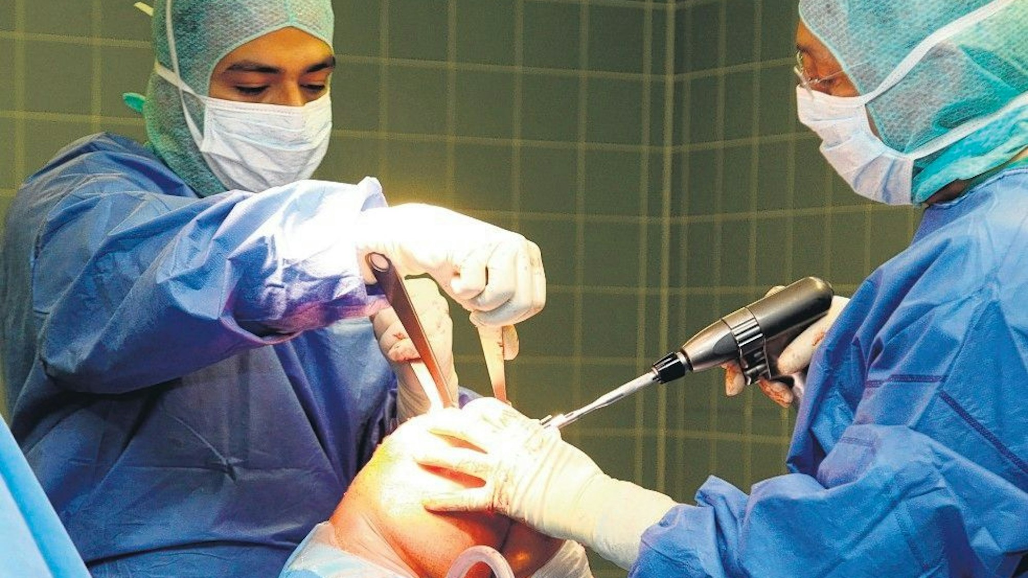 Zwei Ärzte in OP-Kleidung, Masken und Hauben arbeiten an einem Knie.