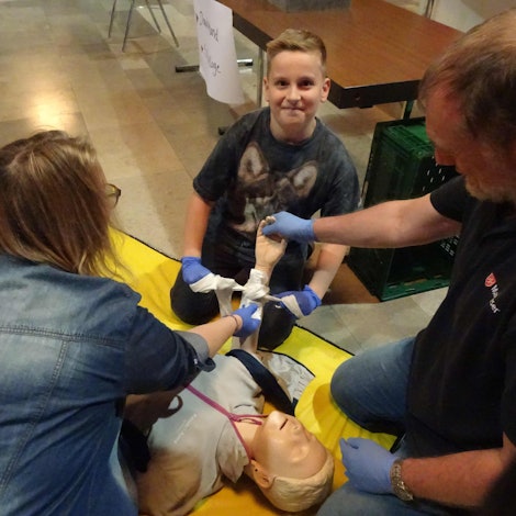 Erste-Hilfe-Ausbilder Jürgen Grenner erklärt dem zehnjährigen Moritz und seiner Mutter Sandra Schwenker aus Rheinbach, wie sie einen Druckverband so anlegen können, dass die Blutung gestoppt wird. Dazu brauchen sie: eine Sterile Wundauflage, ein Dreieckstuch und eine Wundauflage.