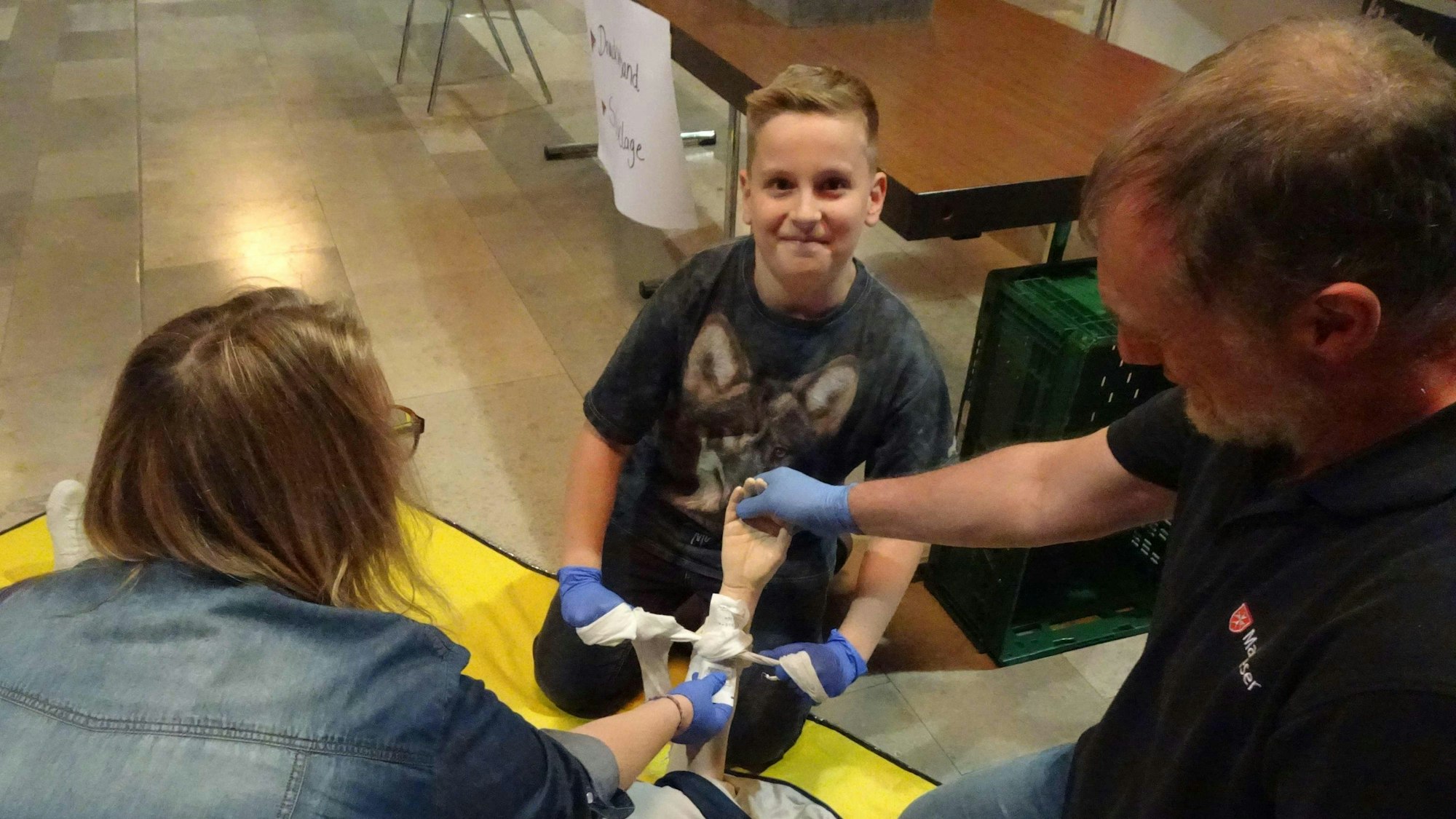 Erste-Hilfe-Ausbilder Jürgen Grenner erklärt dem zehnjährigen Moritz und seiner Mutter Sandra Schwenker aus Rheinbach, wie sie einen Druckverband so anlegen können, dass die Blutung gestoppt wird. Dazu brauchen sie: eine Sterile Wundauflage, ein Dreieckstuch und eine Wundauflage.