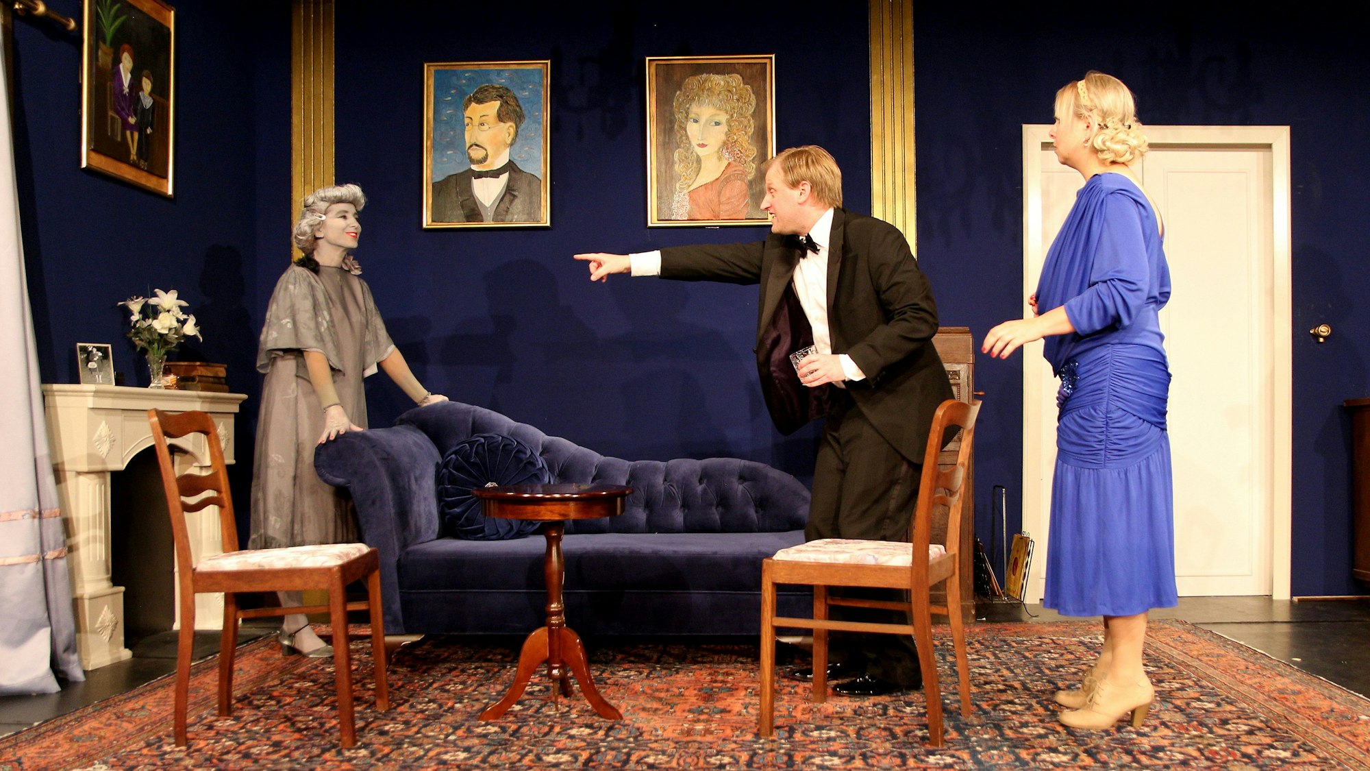 Ein Mann steht zwischen zwei Frauen in einem Wohnzimmer. Er zeigt mit dem Finger in Richtung der Frau links.