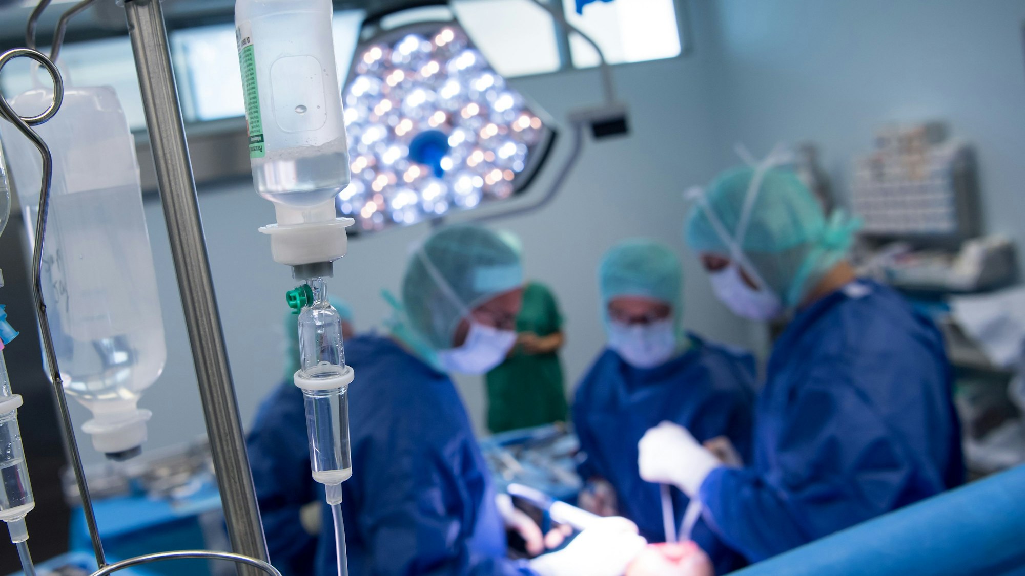 Im Hintergrund wird ein Mensch operiert, im Vordergrund sieht man einen Tropf