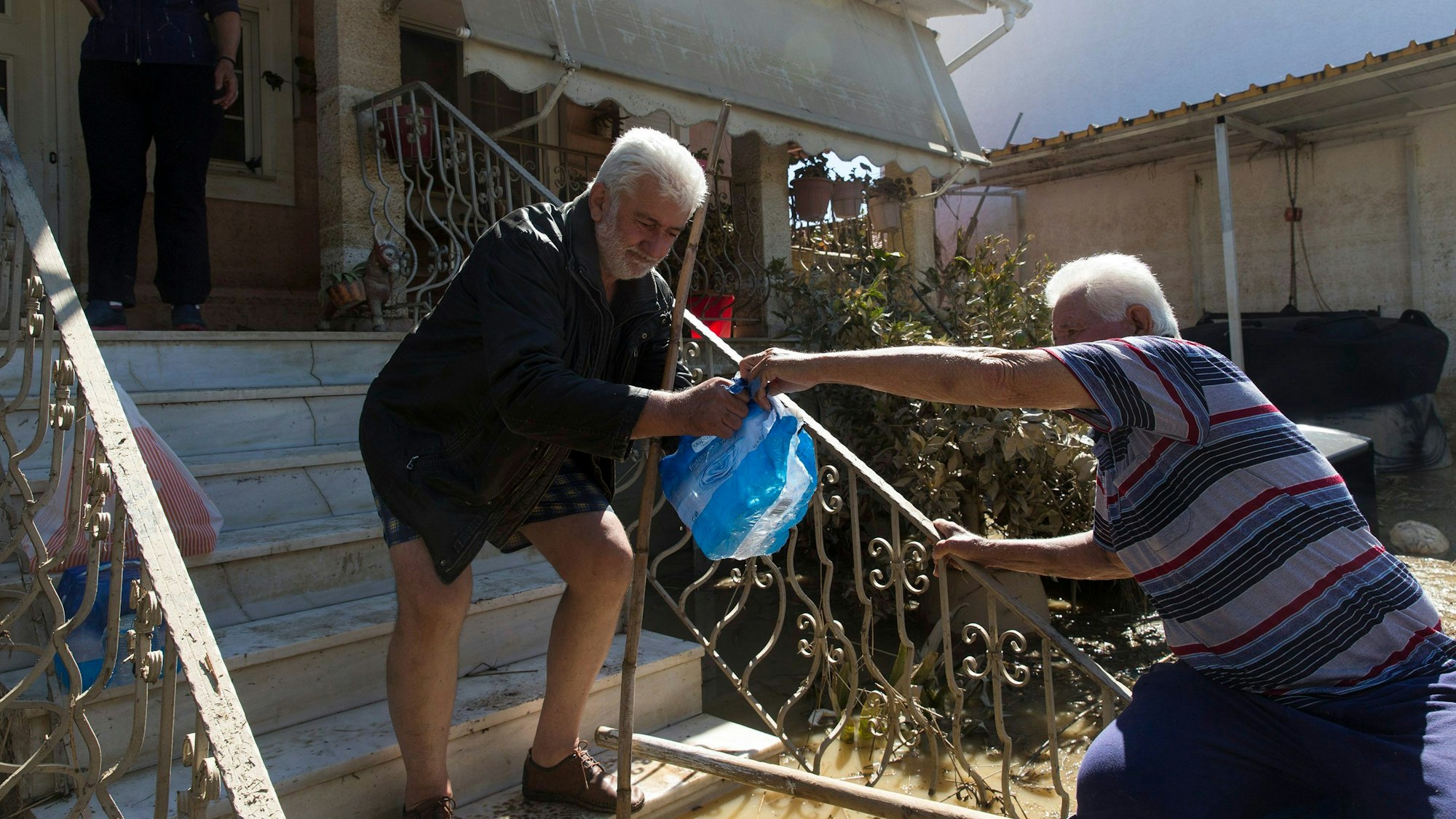 Ein Mann übergibt einem Bewohner eine Tasche mit Vorräten. In den von Überschwemmungen betroffenen Gebieten Mittelgriechenlands sind die Wasserstände weiter gesunken.
