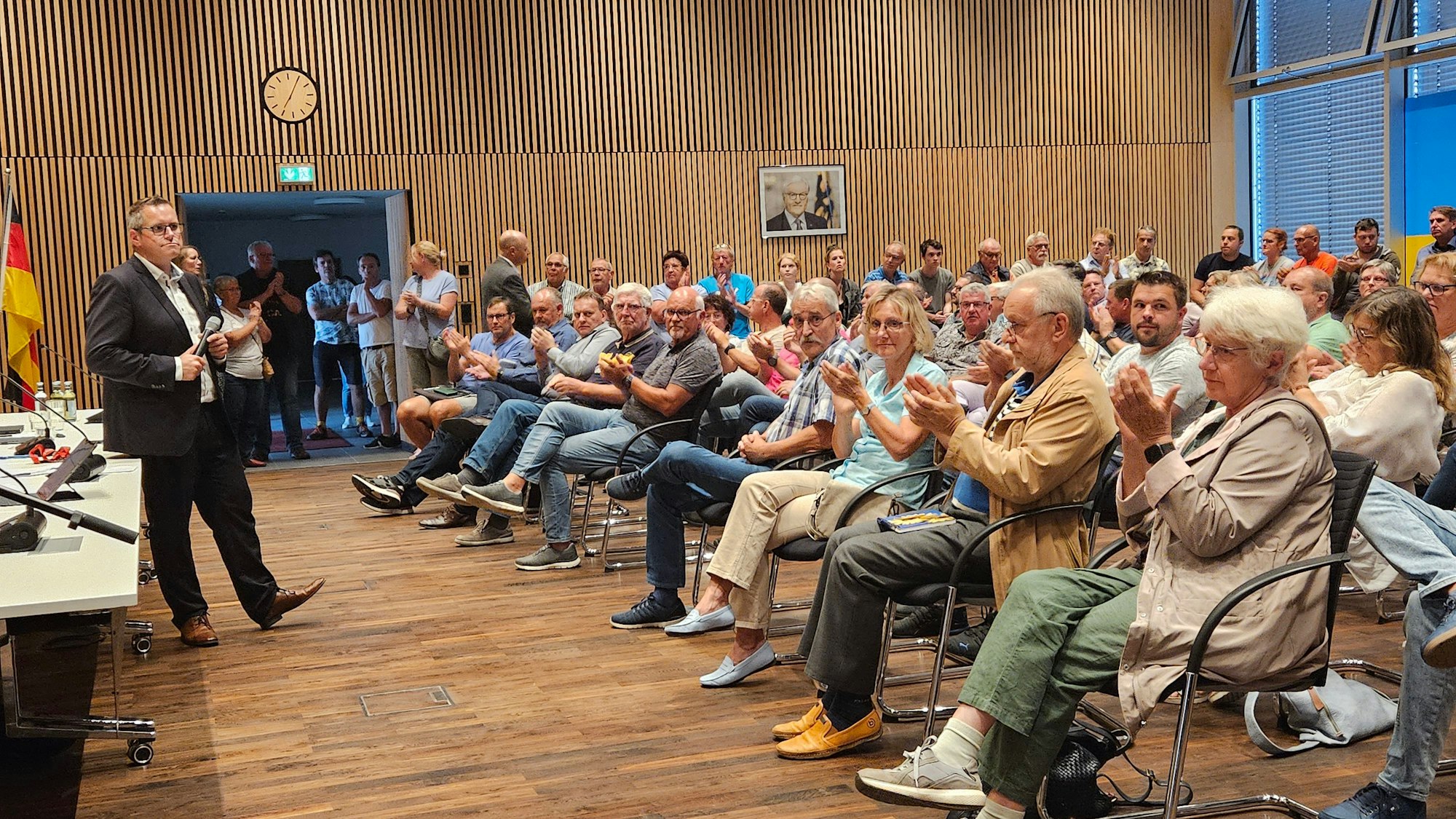 Ein Mann steht in einem großen Saal vor vielen Menschen, die in mehreren Stuhlreihen sitzen und applaudieren.