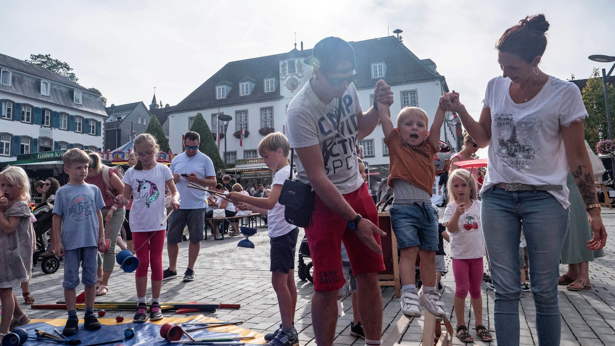 Eine Familie auf dem Marktplatz vor dem Rathaus von Wipperfürth. Mutter und Vater halten ihren Sohn an den Händen, während das Kind über eine Slackline balanciert.