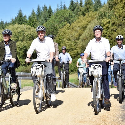 Die Politiker fahren mit dem Rad über einen Weg. Im Hintergrund sind Bäume zu erkennen.