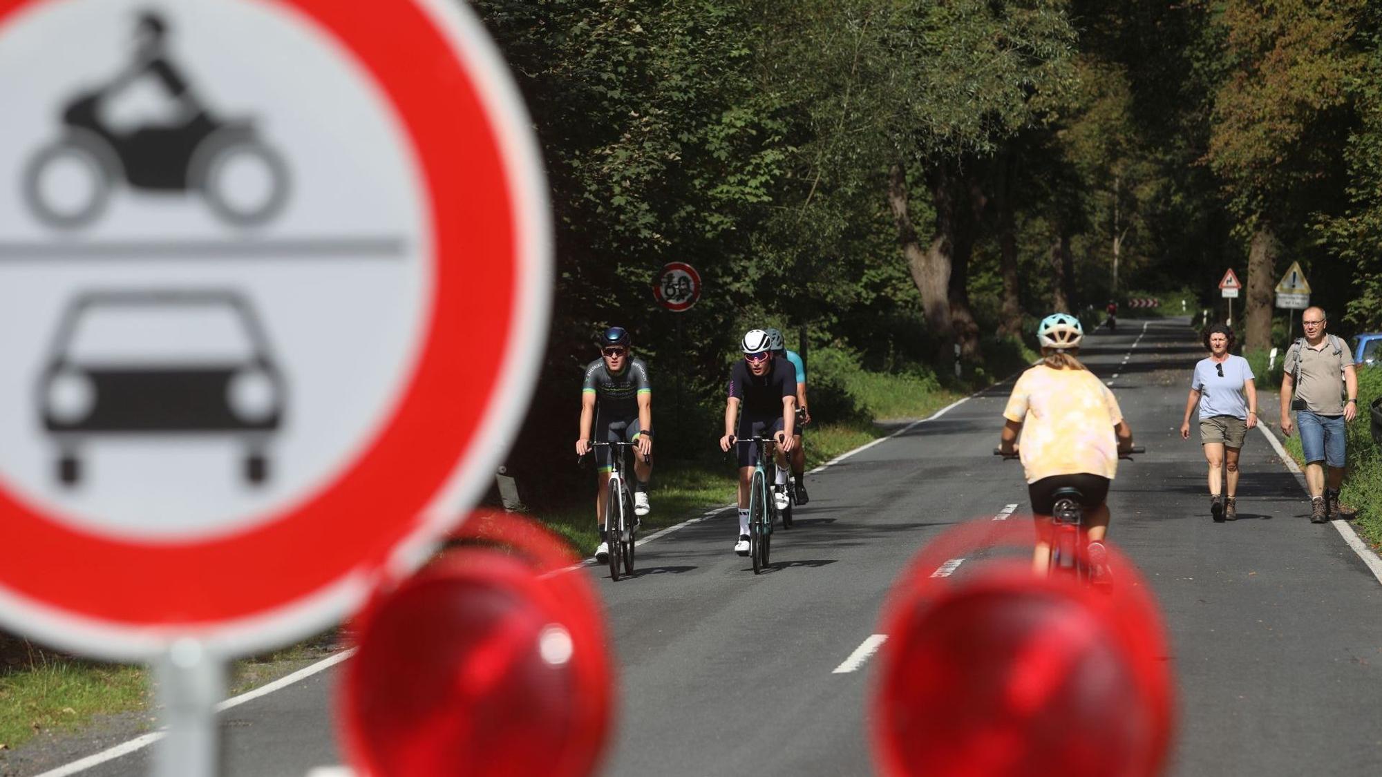 Hinter einem Verbotsschild für Motorräder und Autos sieht man Radfahrer und Wanderer auf einer langen, baumgesäumten Straße.