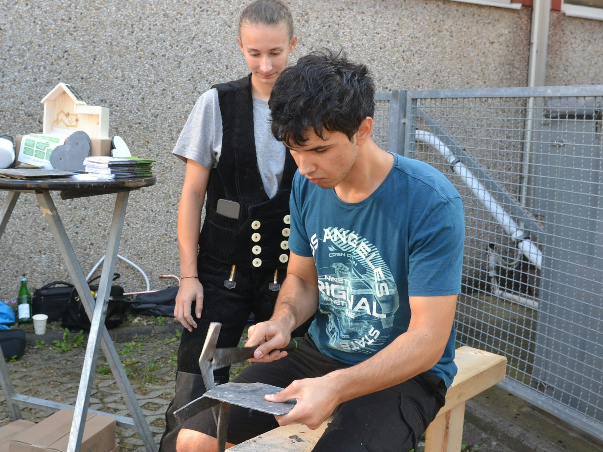 Ein junger Dachdecker steht in Zunftkleidung hinter einem Jugendlichen, der bei der Ausbildungsbörse das Bearbeiten einer Schieferplatte ausprobiert.