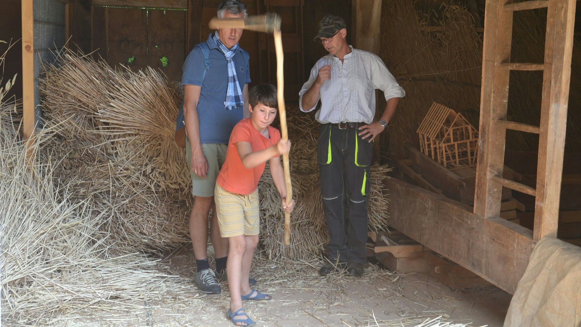 Ein Junge hält eine Sense in die Luft, zwei Männer beobachten ihn dabei.