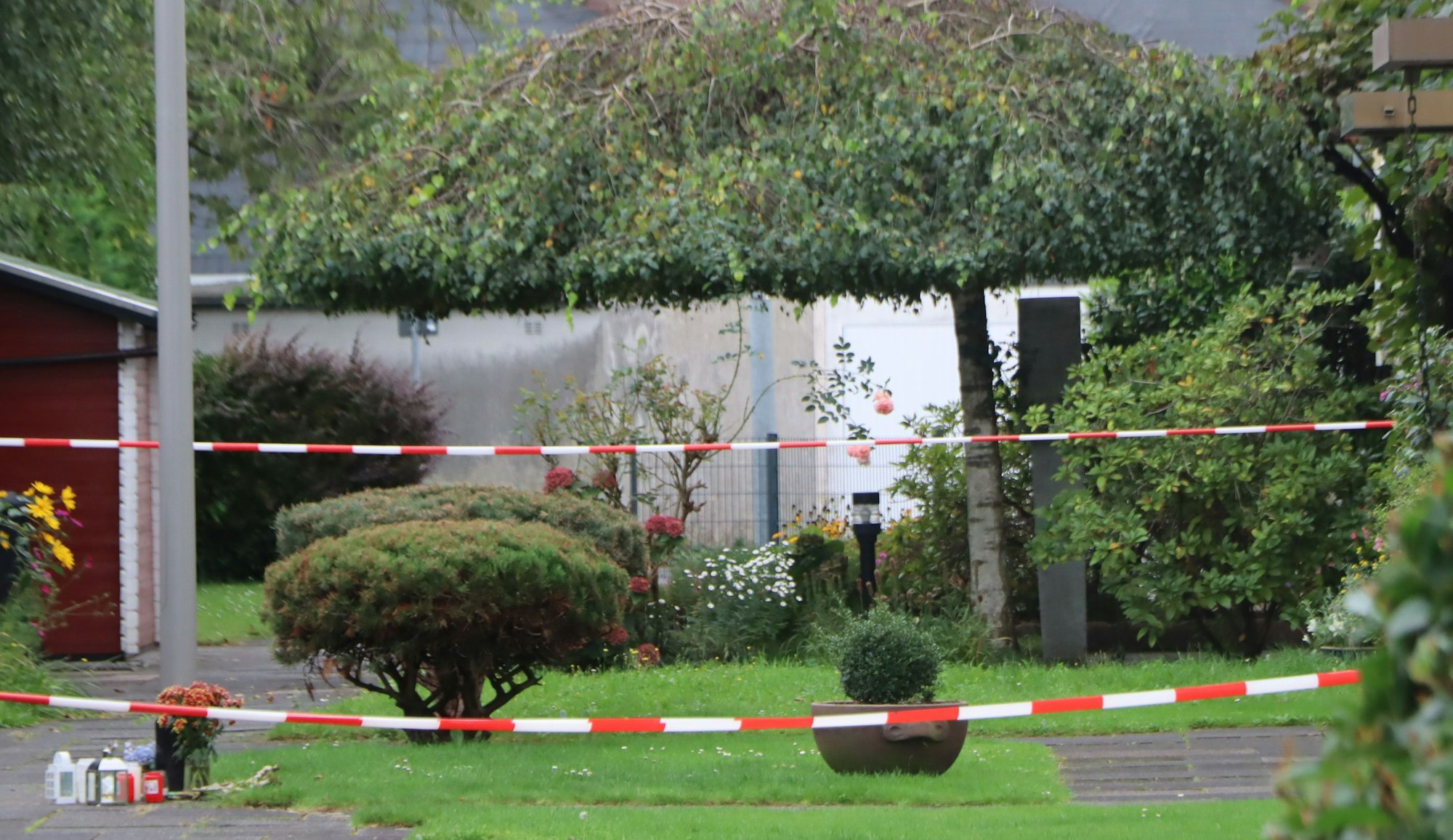 Auf dem Foto ist rot-weißes Flatterband zu sehen, womit das Reihenhaus, in dem der 85-Jährige ermordet worden sein soll, gesichert wird. Dahinter liegen Blumen, die Nachbarn und Freunde niedergelegt haben.