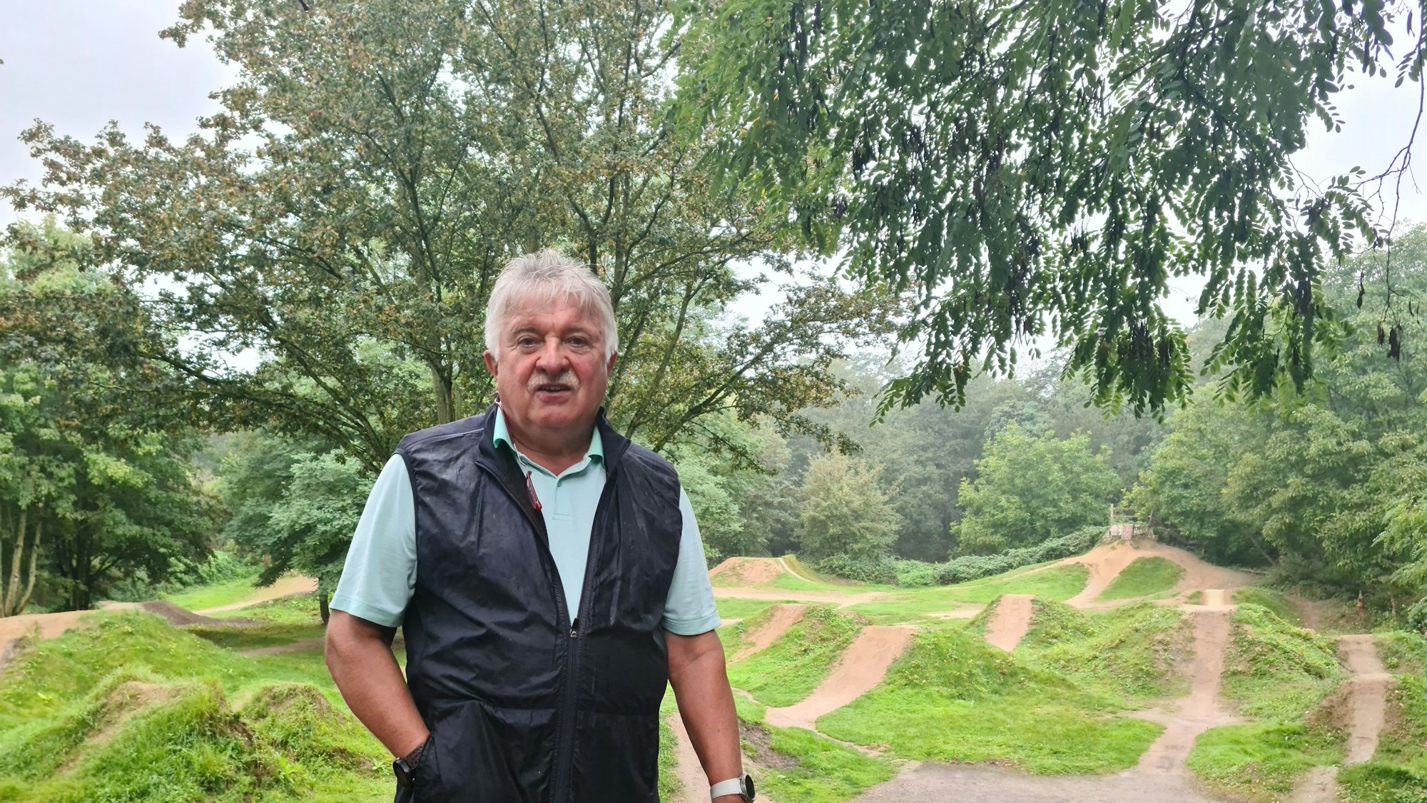 Ein Mann mit grauem Haar und Schnauzbart steht vor hügeligem Gelände.
