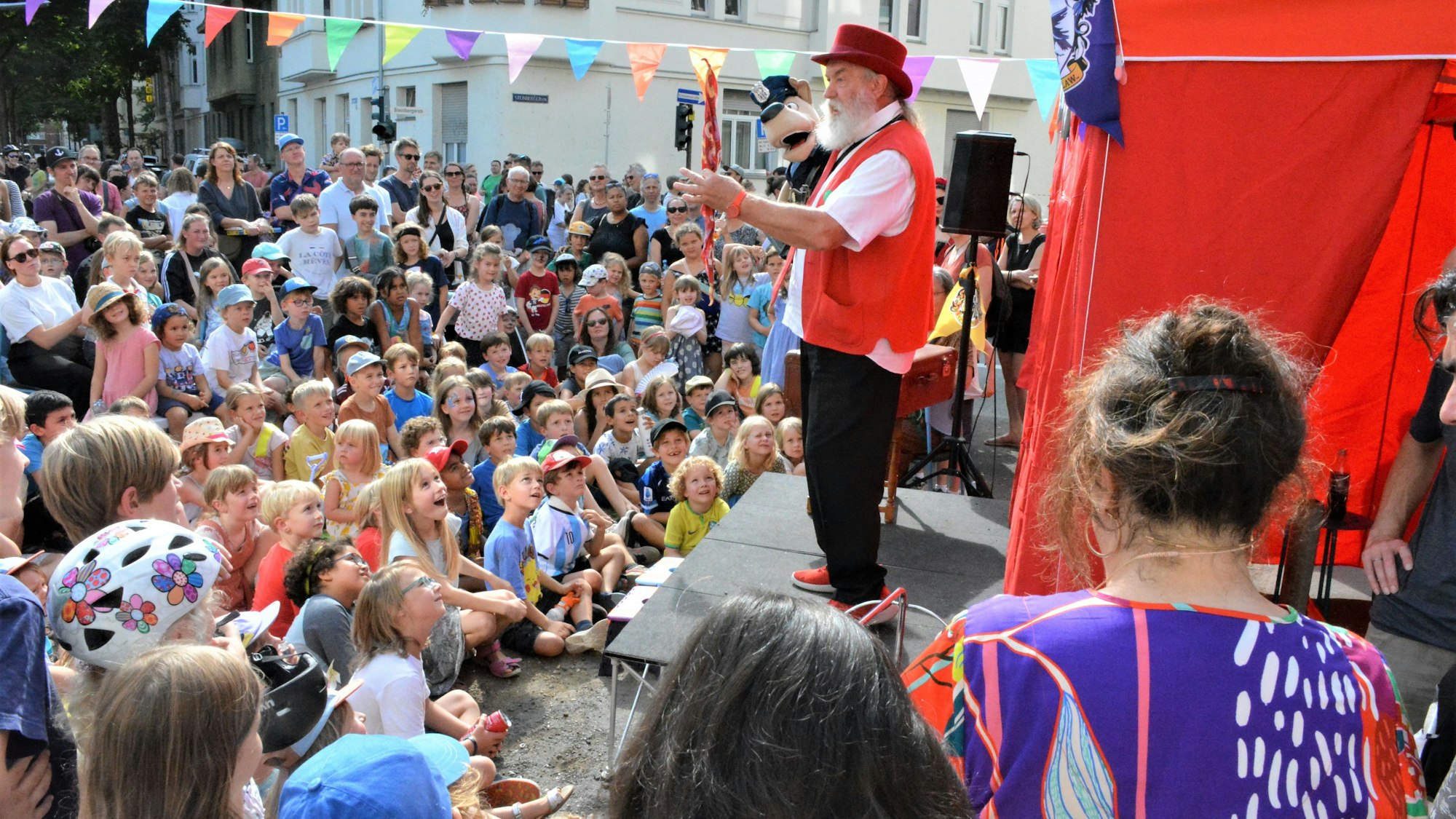 TEin Puppenspieler mit einer Handpuppe steht auf einer Bühne vor zahlreichen Kindern.