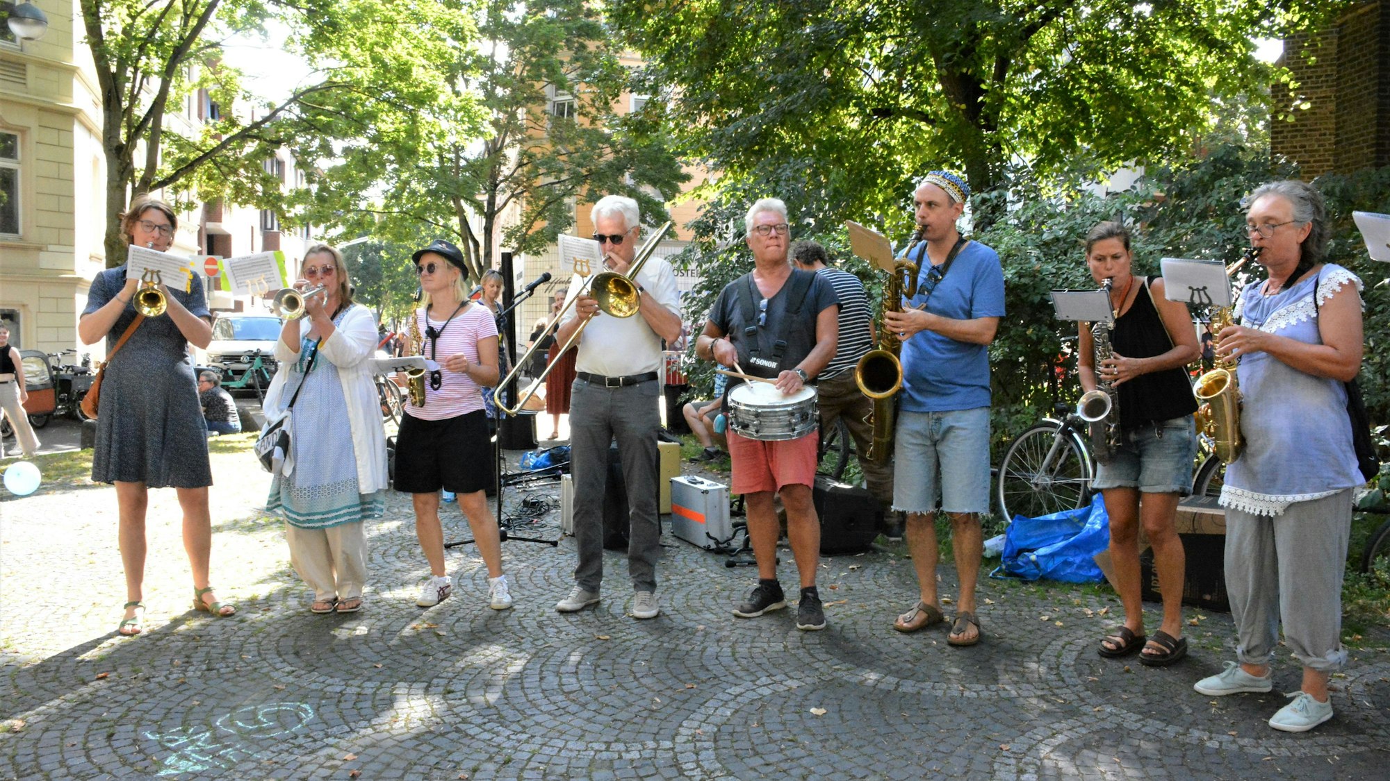 Ein Blaskapelle steht auf einem Platz unter Bäumen und spielt.