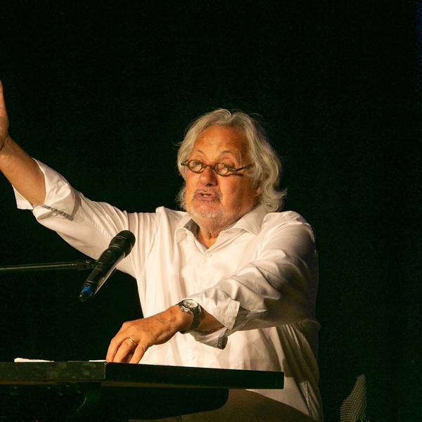 Der Kabarettist Konrad Beikircher sitzt gestikulierend an einem Tisch, vor ihm ein Glas Weißwein.