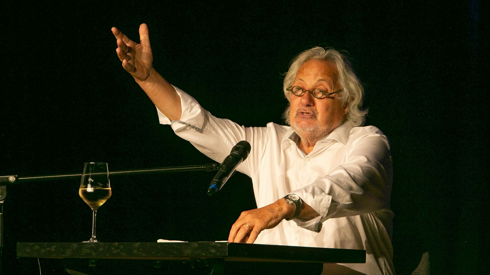 Der Kabarettist Konrad Beikircher sitzt gestikulierend an einem Tisch, vor ihm ein Glas Weißwein.