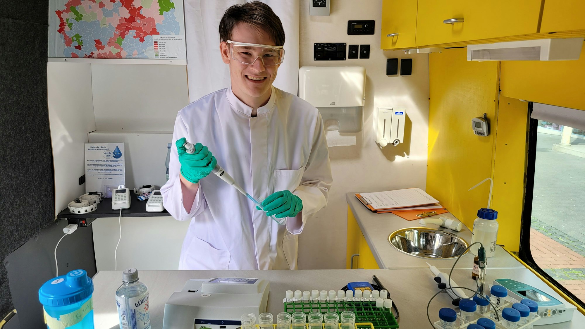 Ein Mitarbeiter nimmt in einem Labor eine Wasserprobe. Auf dem Tisch stehen Flaschen unterschiedlicher Größe.