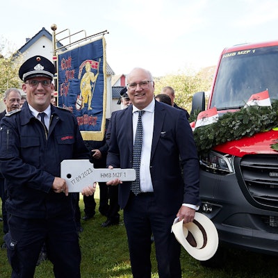 Bürgermeister Jochen Weiler und Löschgruppenführer Bernd Freuen halten den überdimensionalen Fahrzeugschlüssel. Hinter ihnen Feuerwehrleute mit einer Fahne und das zur Übergabe festlich geschmückte neue Fahrzeug.