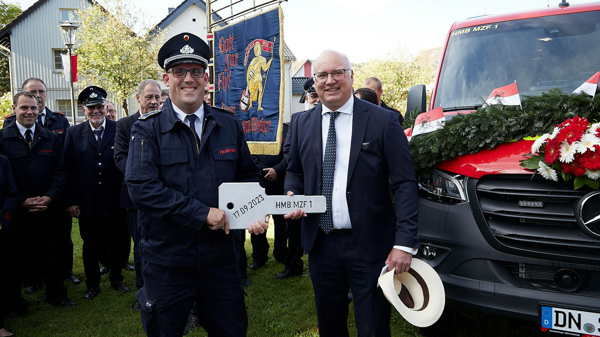 Bürgermeister Jochen Weiler und Löschgruppenführer Bernd Freuen halten den überdimensionalen Fahrzeugschlüssel. Hinter ihnen Feuerwehrleute mit einer Fahne und das zur Übergabe festlich geschmückte neue Fahrzeug.