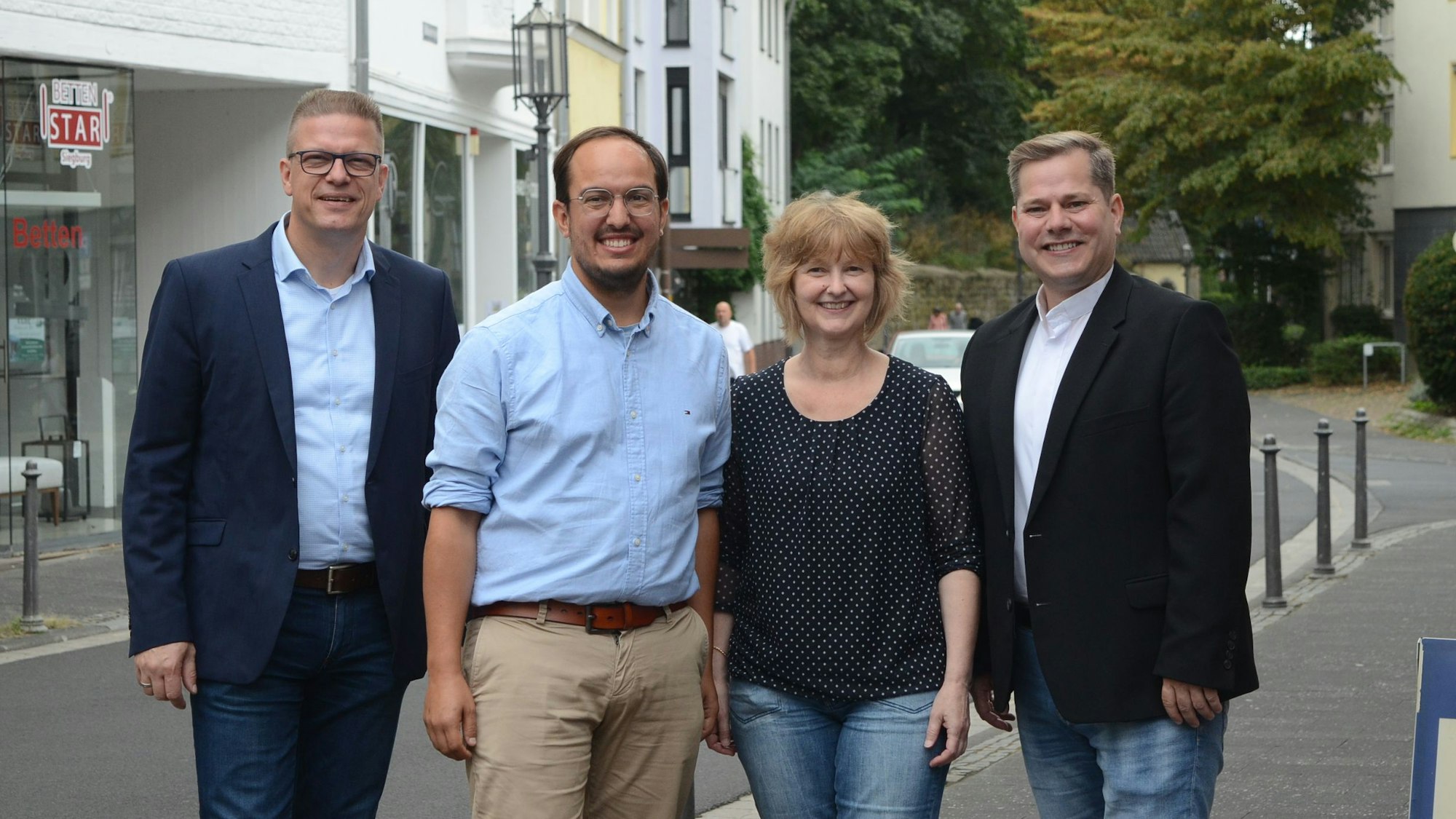 Drei Männer und eine Frau stehen auf dem Gehweg an einer Straße.