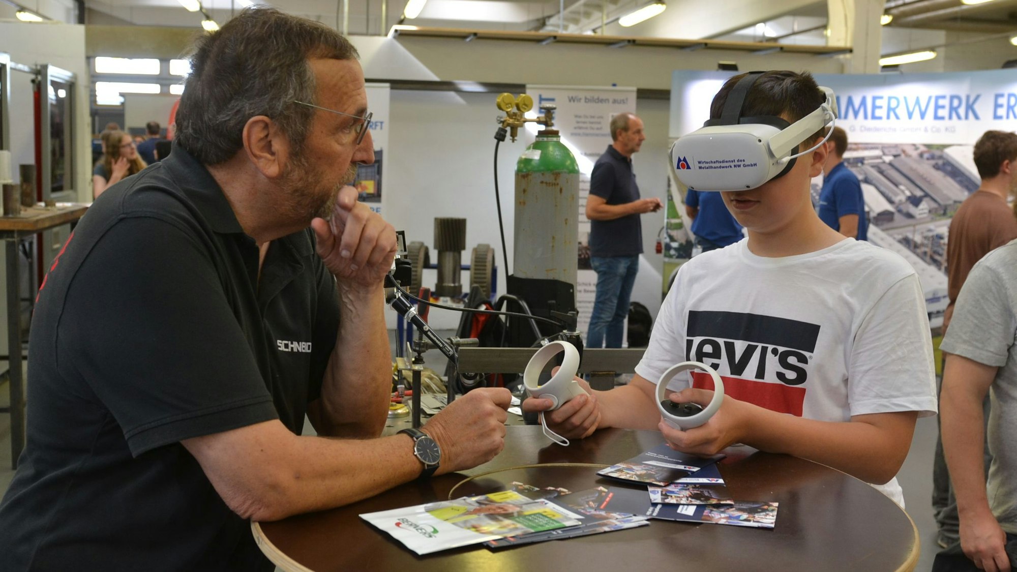 Der Neuntklässler Noah Gerhards trägt eine VR-Brille und hält in jeder Hand eine Bedienkonsole. Metallbaumeister Wilfried Schneider erklärt ihm, wie die Technik in die Ausbildung eingebunden werden kann.