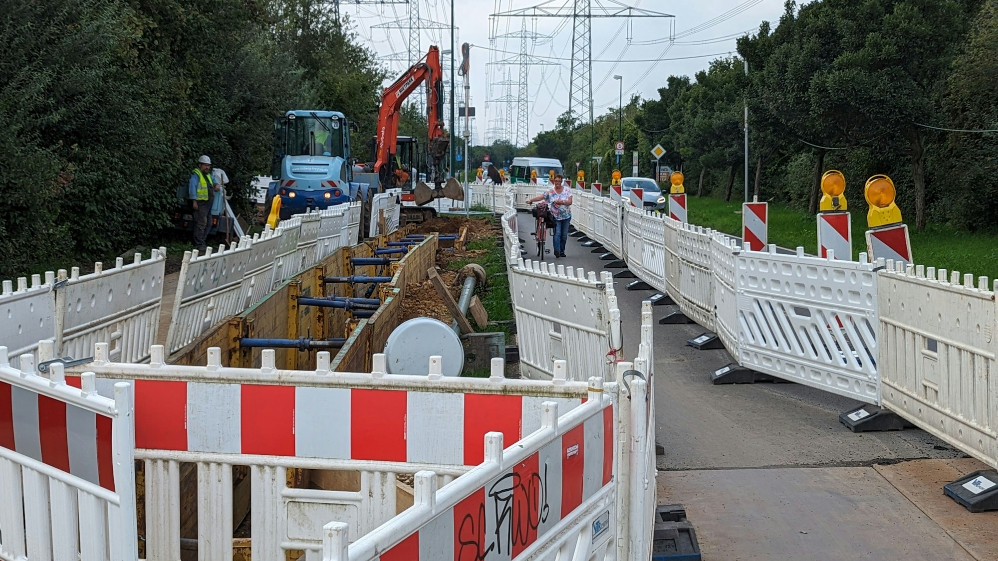 Eine lange Reihe von Baustellenzäunen und ein Bagger an einer Baugrube entlang einer Straße.