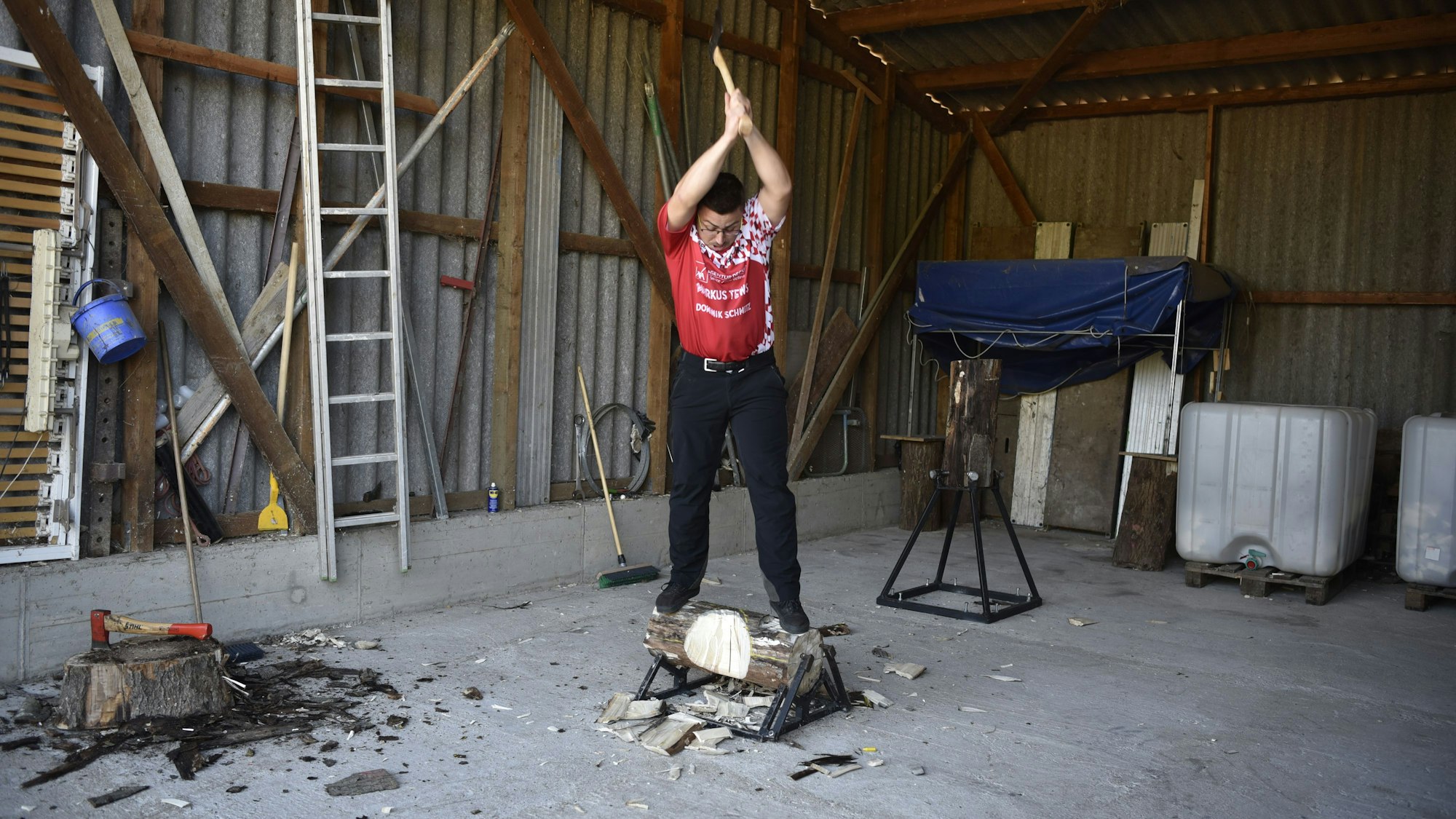 Der Sportholzfäller Leon Pesch steht in einem Schuppen auf einem Baumstamm. Die Axt hält er hoch über dem Kopf, um den Stamm zu zerteilen.