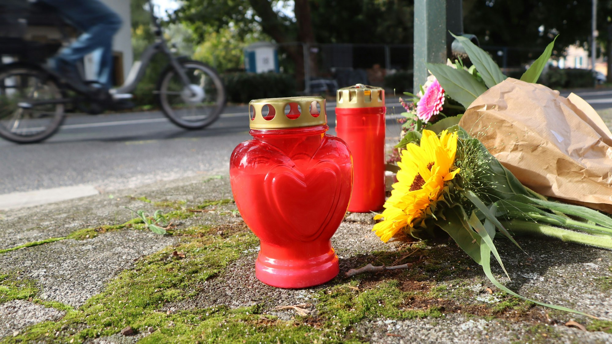 Auf dem Foto sind Blumen und Kerzen an der Unfallstelle in Pulheim zu sehen. Im Hintergrund fährt ein Fahrradfahrer vorbei.