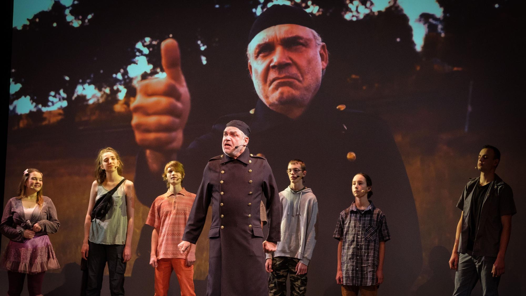 Darsteller eines Theaterstücks stehen vor einer Leinwand, auf der der Schauspieler in der Mitte noch einmal groß zu sehen ist.
