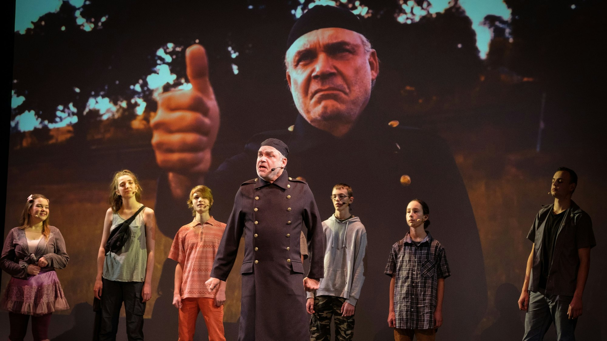 Darsteller eines Theaterstücks stehen vor einer Leinwand, auf der der Schauspieler in der Mitte noch einmal groß zu sehen ist.