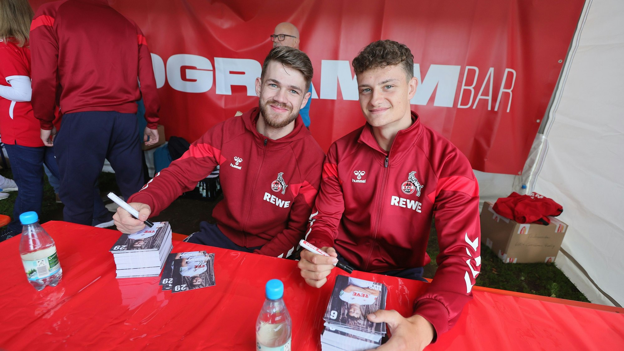 Jan Thielmann und Eric Martel sitzen an einem Tisch und unterschreiben Autogrammkarten.