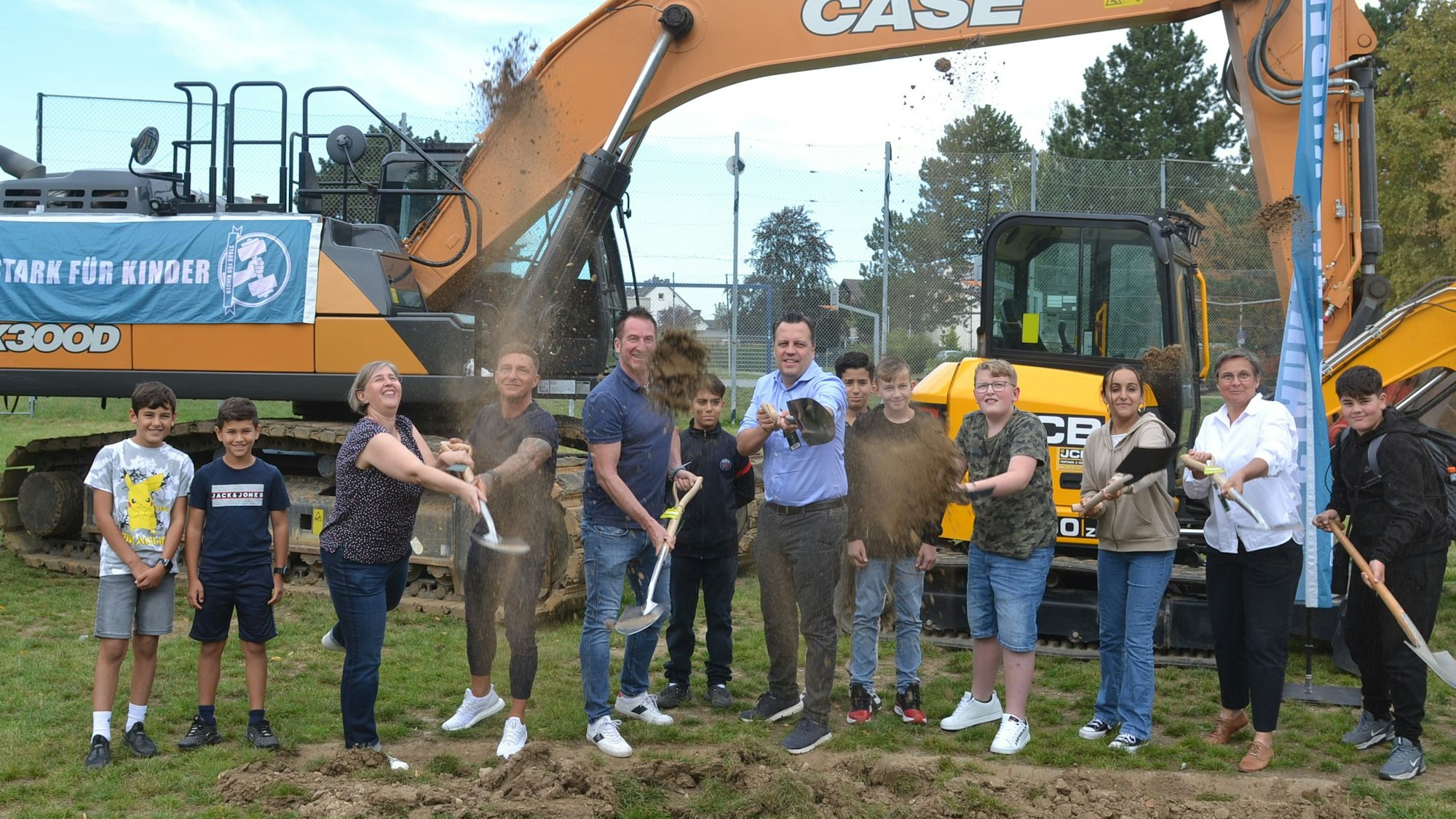 Eine Gruppe Menschen steht vor einem Bagger auf einer Wiese. Zu einem symbolischen ersten Spatenstich schaufeln sie ein wenig Erde.
