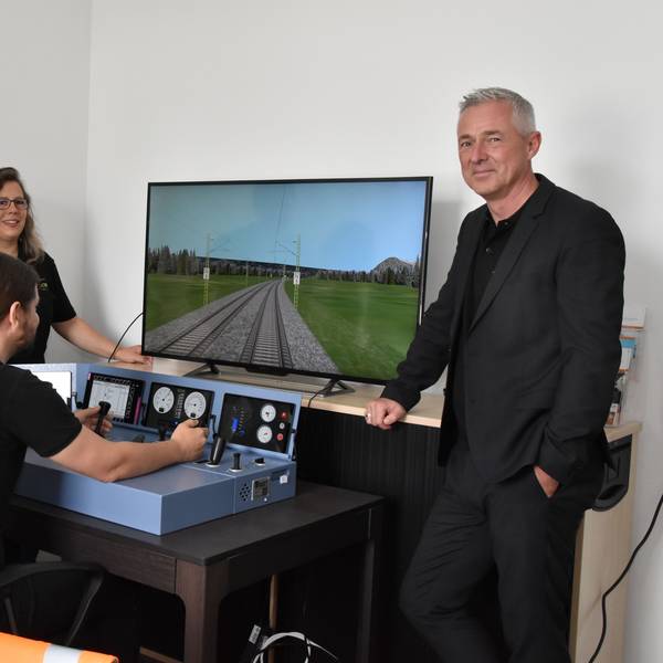 Vier Personen stehen am Simulator eines Lok-Führerstandes. Einer von ihnen steuert die Lok über eine Strecke, die auf dem Bildschirm zu sehen ist.