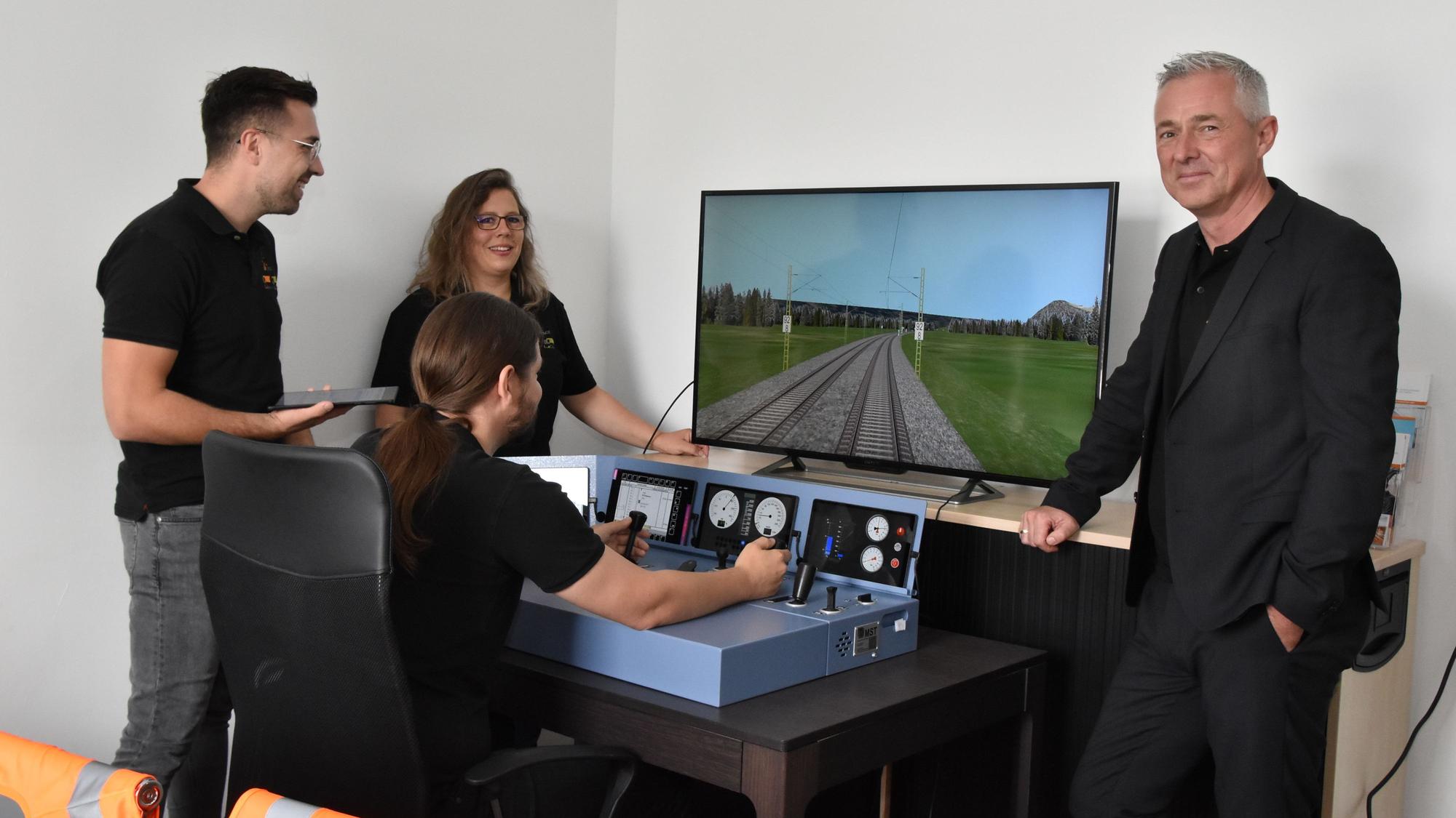 Vier Personen stehen am Simulator eines Lok-Führerstandes. Einer von ihnen steuert die Lok über eine Strecke, die auf dem Bildschirm zu sehen ist.