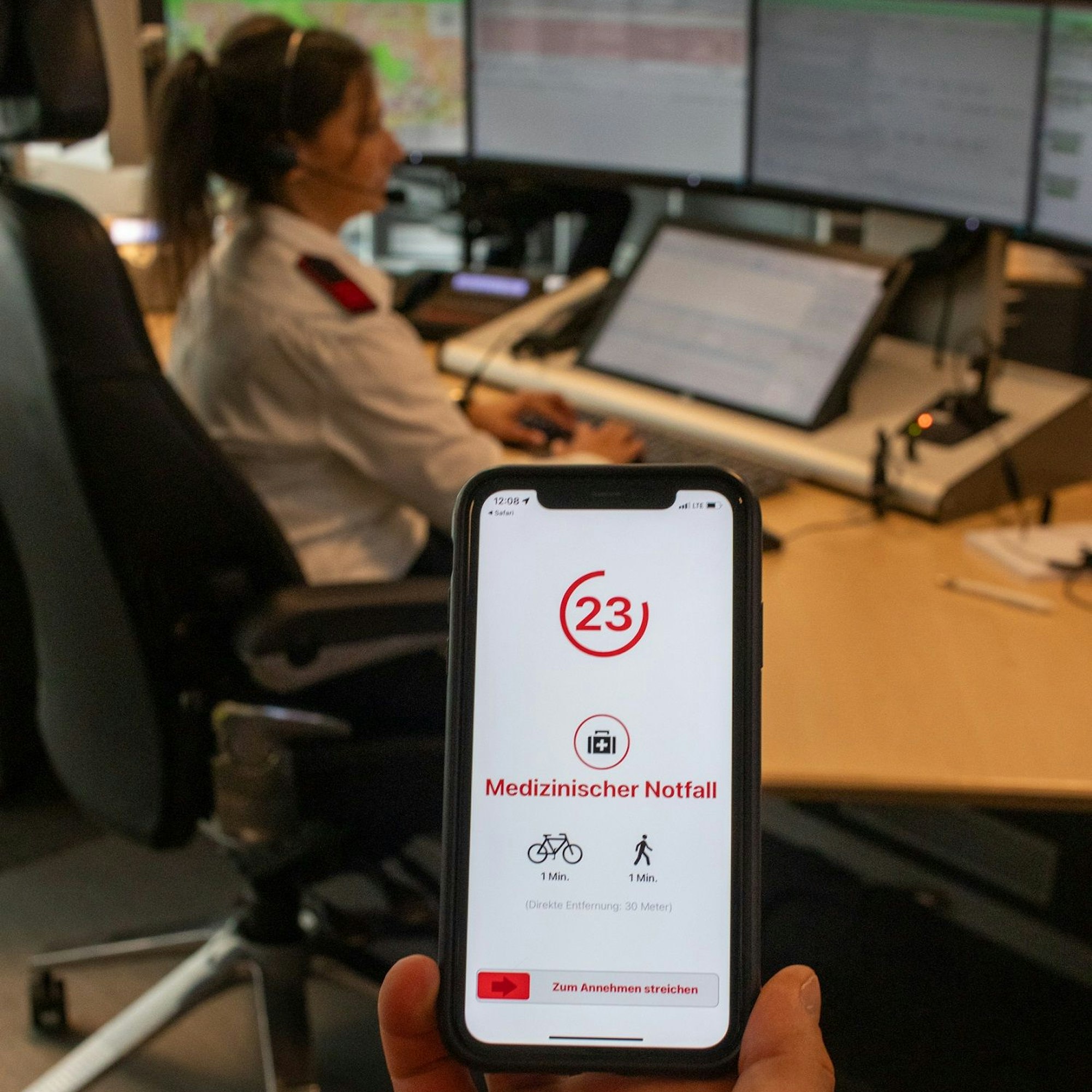 Eine Hand hält ein Handy mit Katretter-App vor eine Arbeitsplatz der Feuer- und Rettungsleitstelle des Rheinisch-Bergischen Kreises.