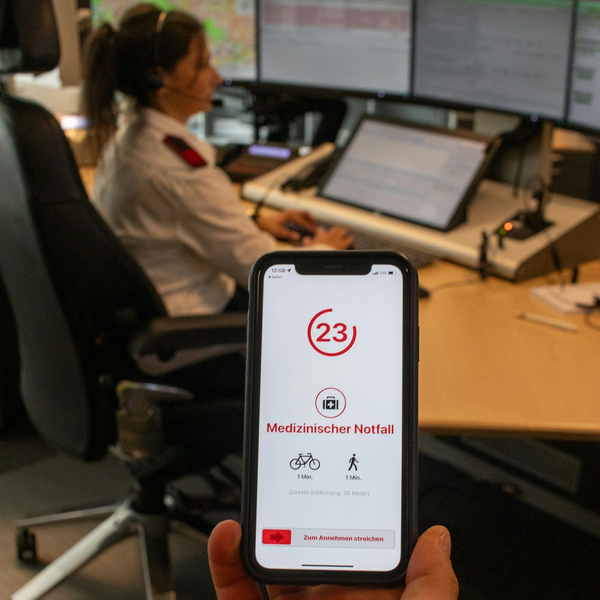 Auf einem Handy-Bildschirm ist die App Katretter zu sehen. Im Hintergrund ist unscharf ein Arbeitsplatz der Feuer- und Rettungsleitstelle des Rheinisch-Bergischen Kreises zu sehen.