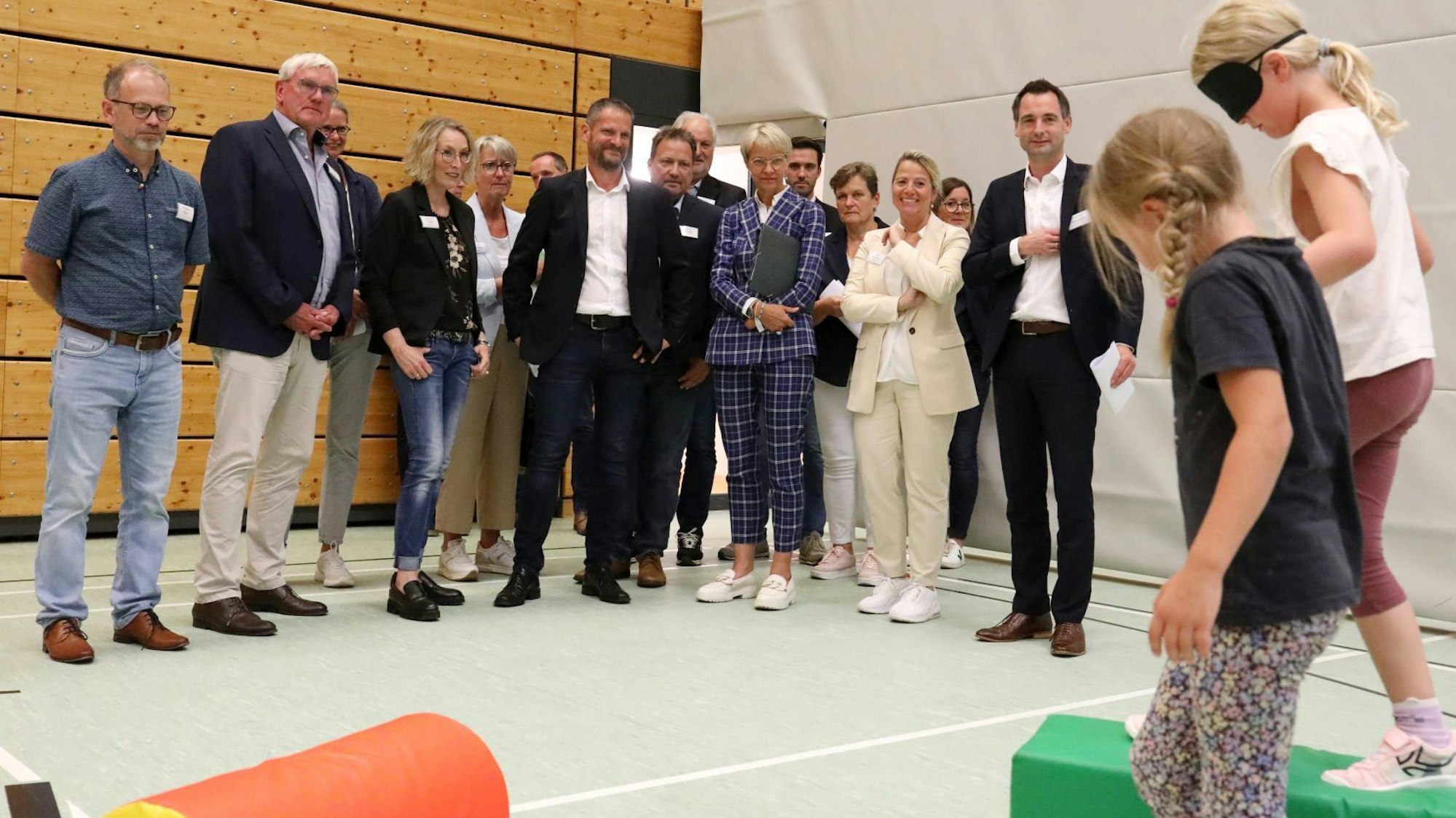 Eine Gruppe von Menschen schaut zwei Schülerinnen bei einer Übung zu: Eines der Kinder führt eine Mitschülerin, deren Augen verbunden sind, über einen kleinen Hindernisparcours.
