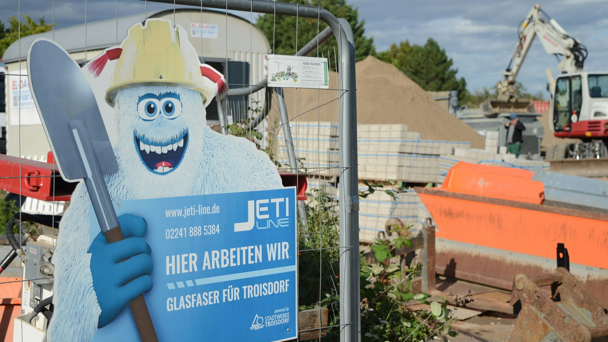 Ein blaues Baustellenschild mit einem Yeti ist vor einem Bauzaun einer Großbaustelle zu sehen.