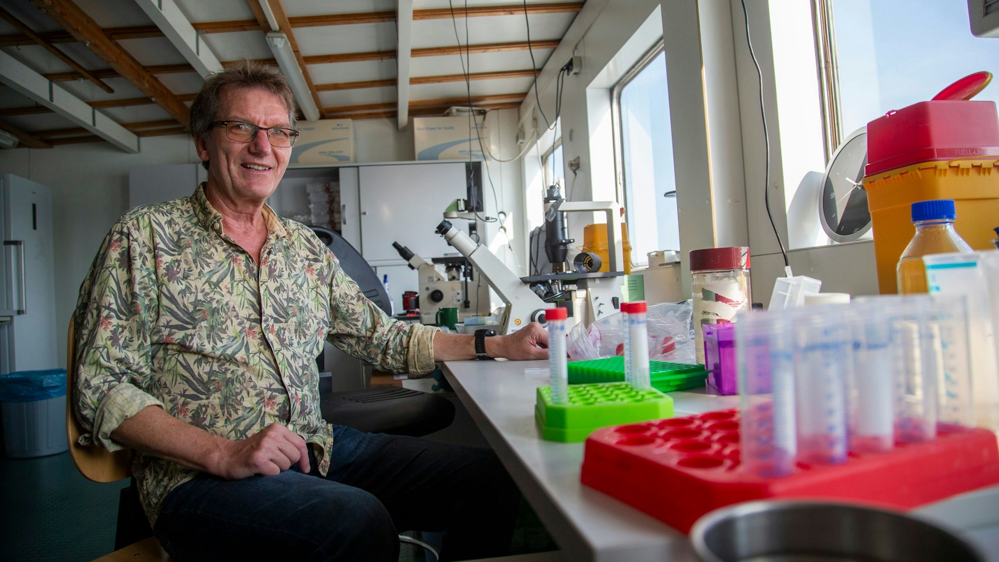 Ein Mann sitzt an einem Tisch, auf dem Mikroskope und Röhrchen stehen.